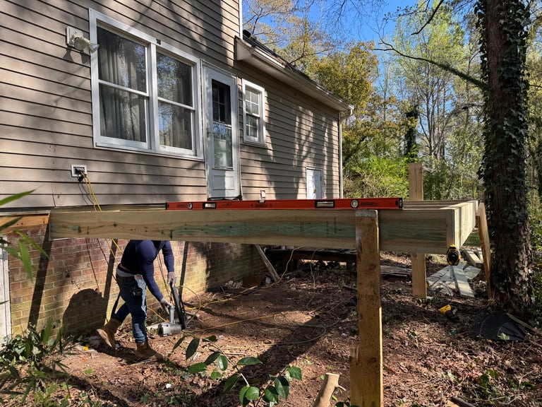 Person working under a wooden deck structure attached to a gray house with trees in background