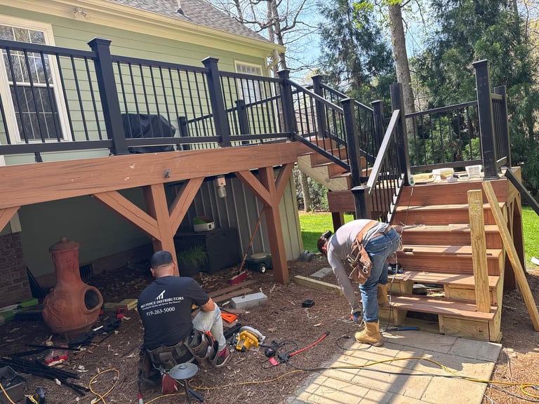 Person building wooden deck stairs under an elevated house deck with black railings and yellow siding