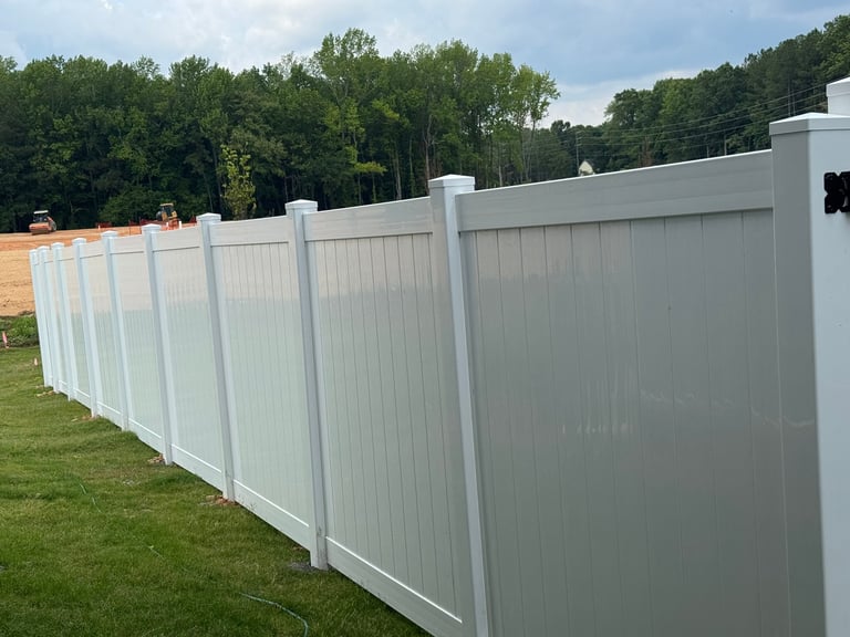 Long white vinyl fence installed along green grass with trees and construction area in background